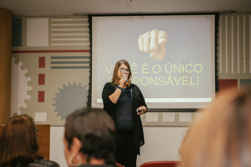 Priscilla Cruz em pé no palco, segurando microfone, durante palestra com slide ao fundo que diz “Você é o único responsável!”, em auditório com público presente.