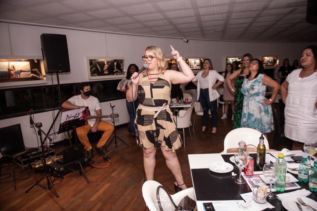 Priscilla Cruz segurando um microfone, sorrindo com confiança durante uma palestra, transmitindo segurança, clareza e autenticidade, com fundo colorido e vibrante.