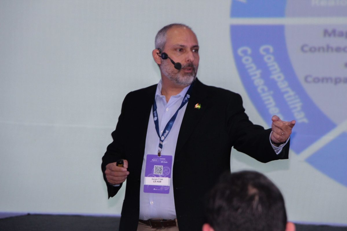 Sergio Frias durante palestra, usando microfone auricular, com crachá da CX HUB, gesticulando em frente a uma apresentação sobre neurociência do consumidor.