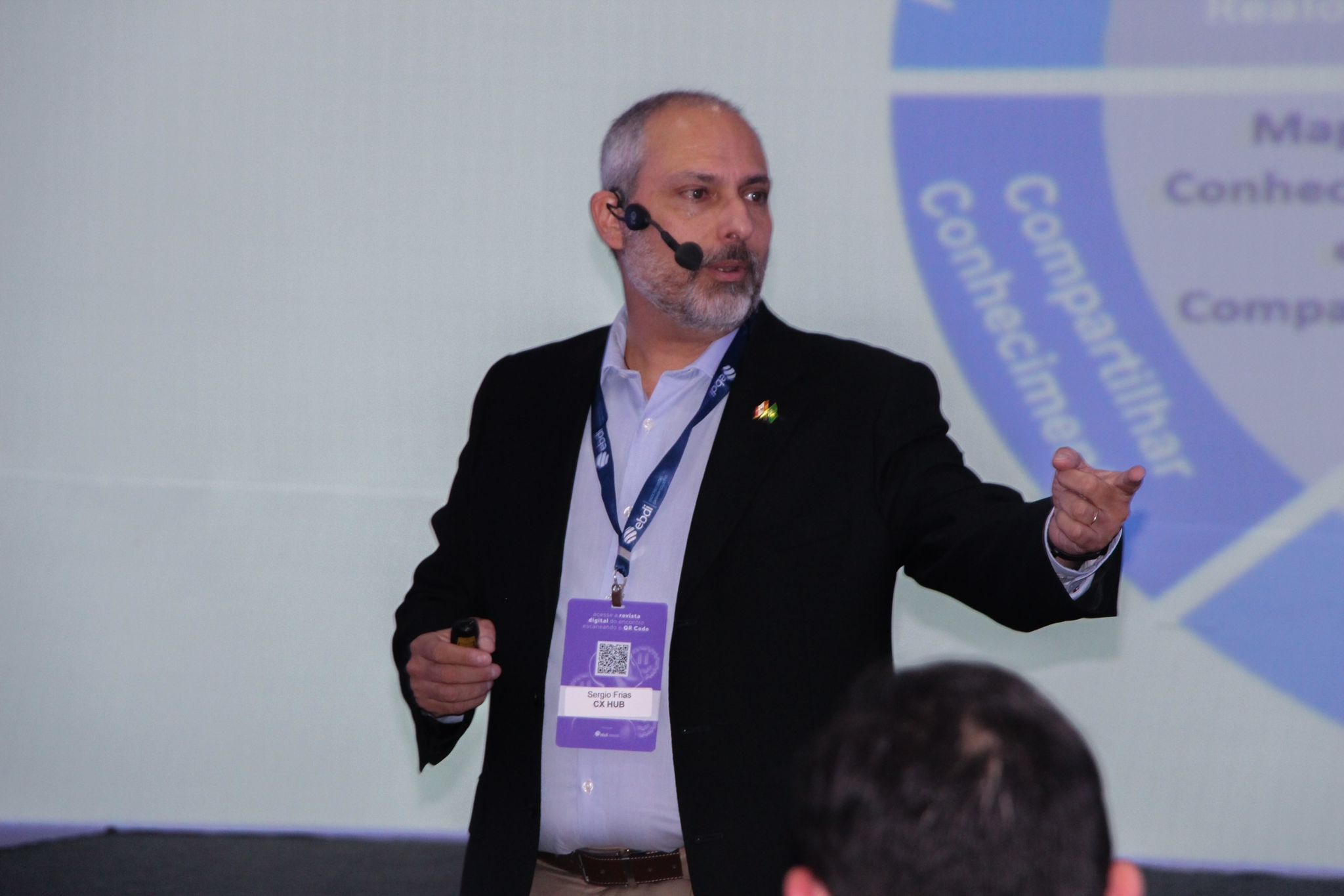 Sergio Frias durante palestra, usando microfone auricular, com crachá da CX HUB, gesticulando em frente a uma apresentação sobre neurociência do consumidor.
