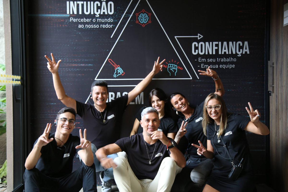 Equipe sorrindo ao lado de Ivan Moré, em frente a um painel com os pilares “Intuição, Confiança e Coragem”, representando os fundamentos da Desobediência Produtiva.