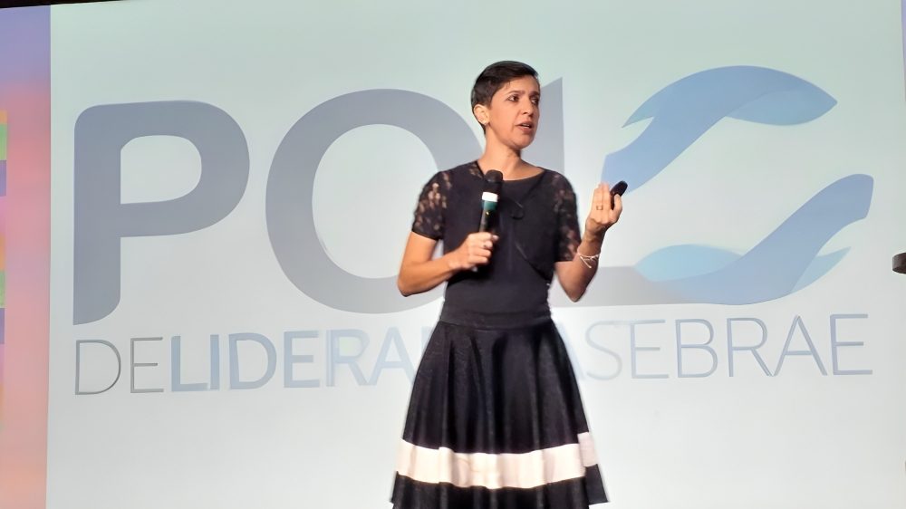 Palestrante em foco durante evento do Sebrae, segurando microfone e apontador, vestida com elegância e expressão engajada.