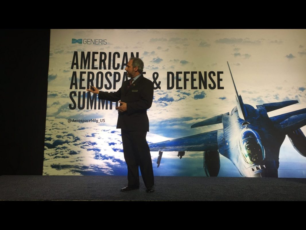 Homem de terno se apresenta em um palco durante o evento American Aerospace & Defense Summit, com imagem de jato de combate ao fundo.