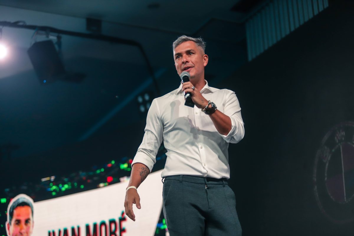 Ivan Moré segurando microfone durante palestra em auditório, usando camisa branca e calça social, com expressão confiante.