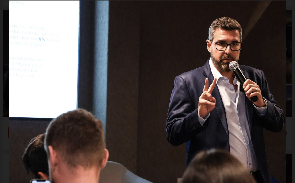 Floriano Schneider em palestra, segurando microfone e fazendo gesto com dois dedos, diante de público atento.
