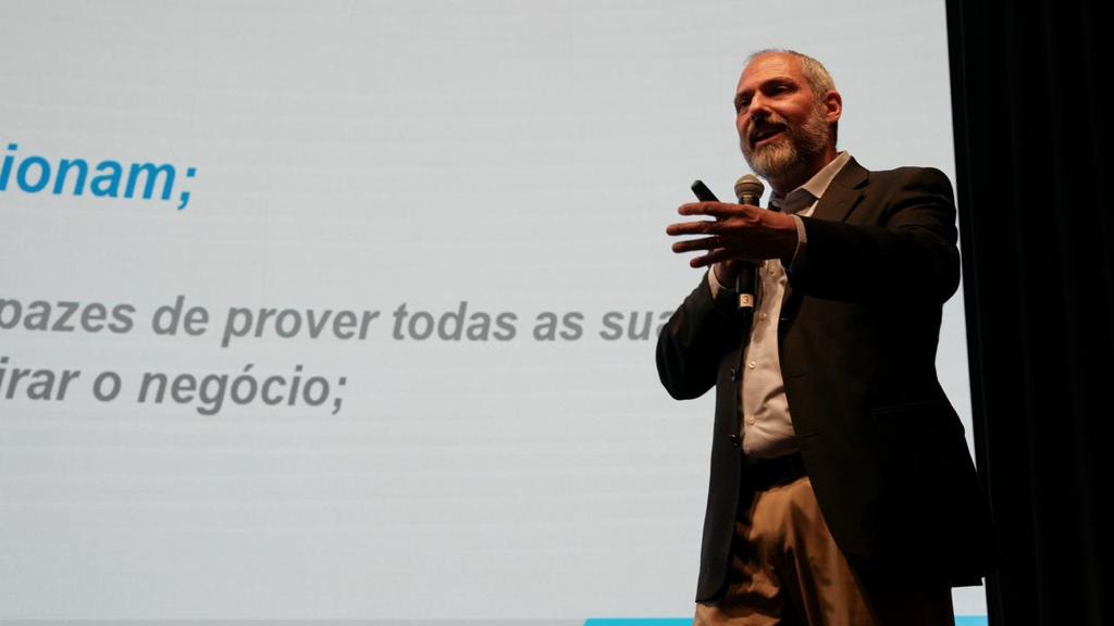 Palestrante de barba grisalha, usando terno escuro e camisa clara, segura um microfone enquanto fala no palco em frente a uma tela de projeção com texto exibido.