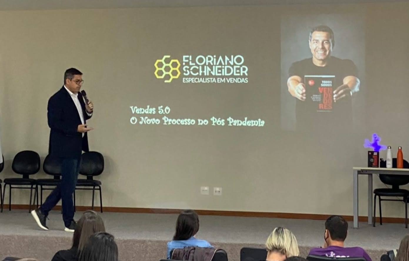 Palestrante Floriano Schneider em auditório apresentando o tema “Vendas 5.0 – O Novo Processo no Pós-Pandemia”, com projeção de seu nome, título de especialista em vendas e imagem segurando o livro Todos Somos Vendedores.