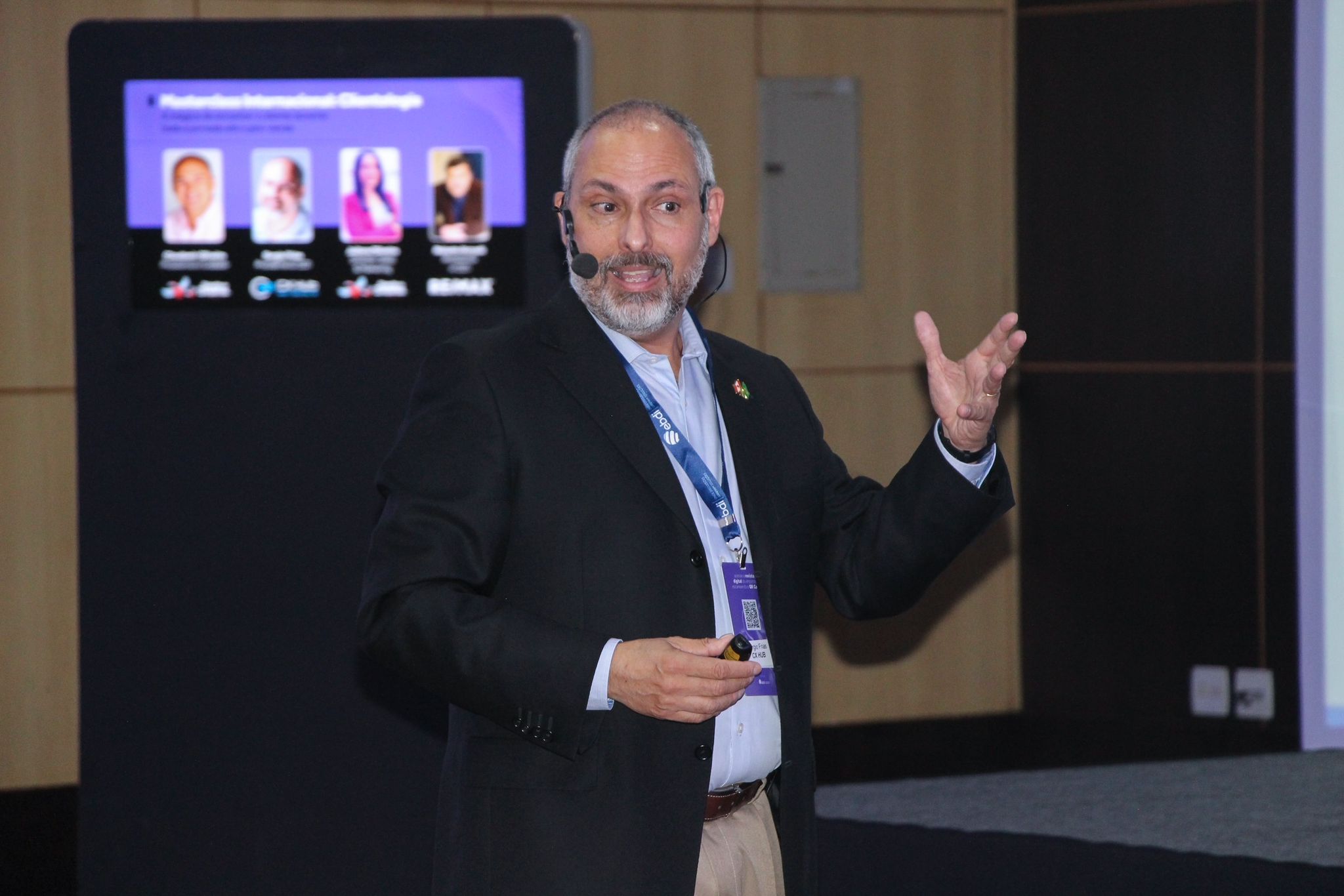 Homem de terno cinza escuro e camisa azul clara gesticulando com a mão direita em um palco durante uma apresentação, com uma tela preta exibindo quatro fotos de pessoas atrás dele.