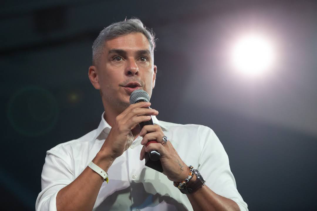 Homem de cabelos grisalhos segurando um microfone com as duas mãos, usando camisa branca e pulseiras, falando em um palco com luz intensa ao fundo.