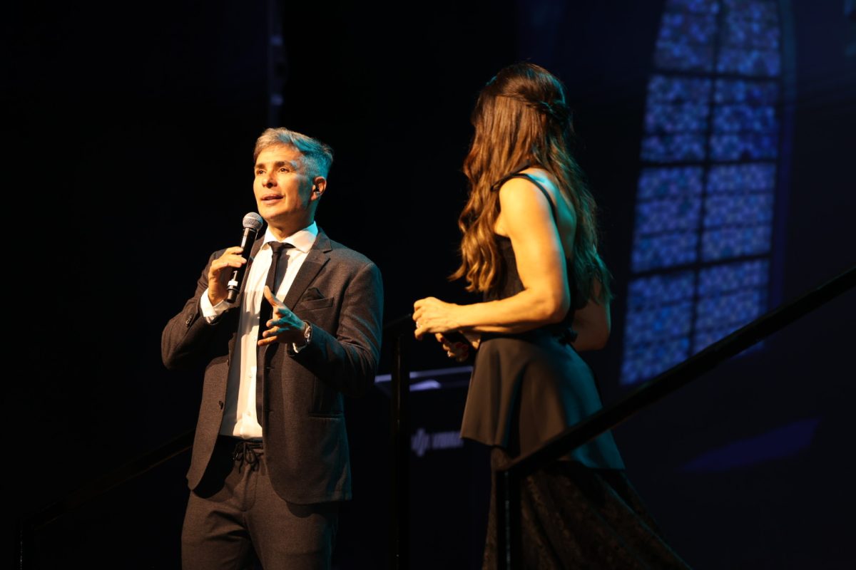Um homem de terno cinza fala ao microfone em um palco iluminado, enquanto uma mulher de vestido preto o escuta atentamente. O ambiente tem iluminação cênica em tons quentes e frios, com fundo escuro e vitrais ao fundo, sugerindo um evento elegante.