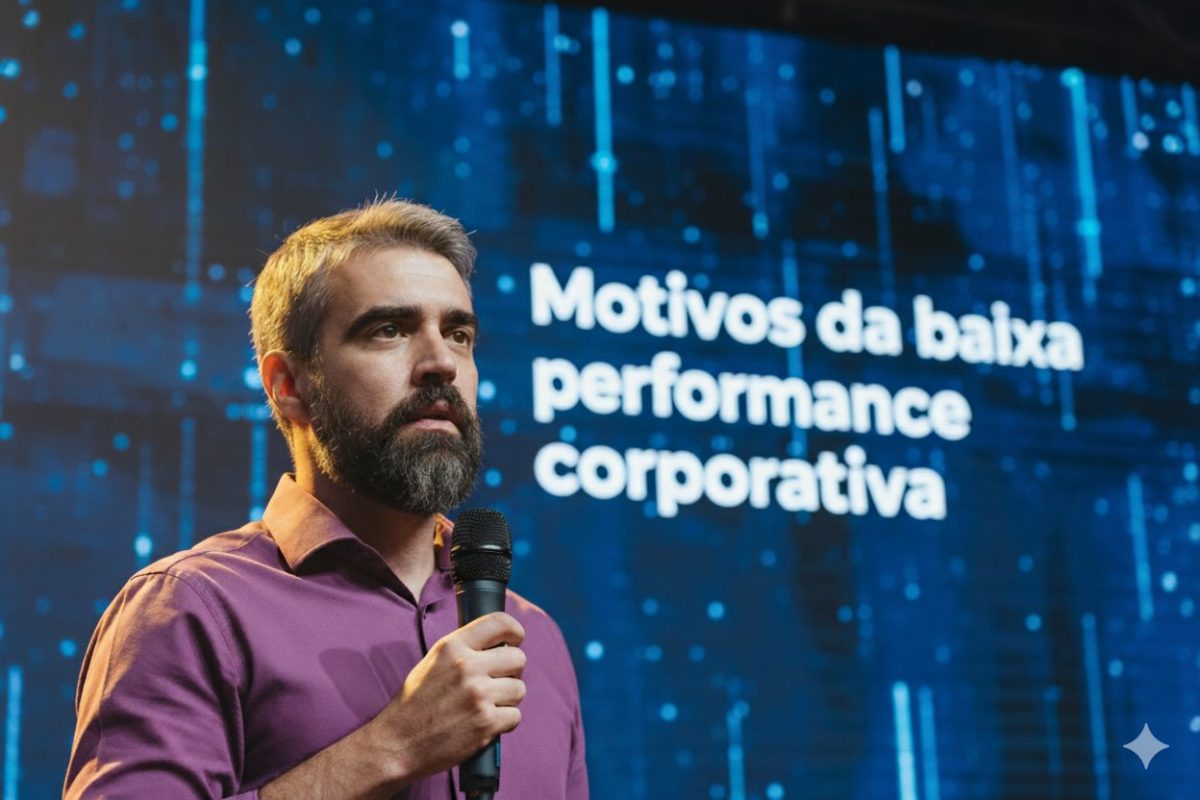 "Um homem com barba e cabelos grisalhos, vestindo uma camisa roxa, está no palco segurando um microfone e olhando para o lado. Ao fundo, um telão azul exibe o texto 'Motivos da baixa performance corporativa'."