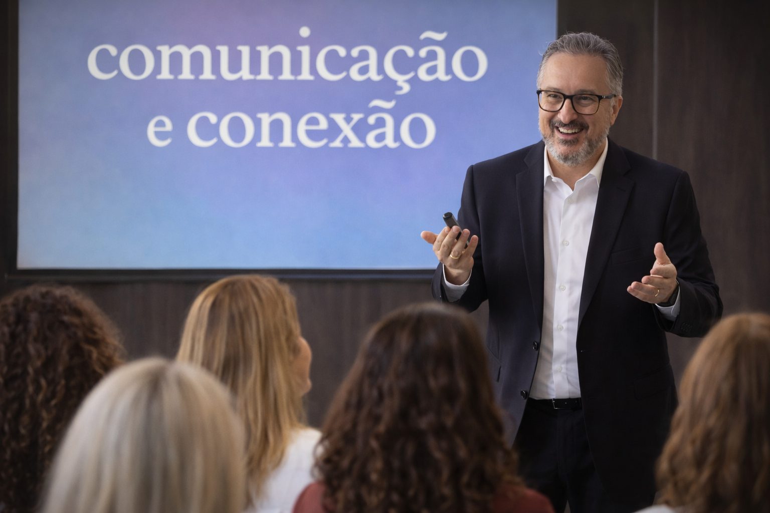 Edson De Paula sorrindo enquanto palestra para um grupo seleto de mulheres diversas, com um telão ao fundo exibindo a frase “comunicação e conexão”, em ambiente corporativo.